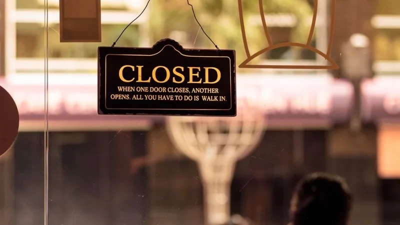A hand-lettered CLOSED sign hanging inside a shop's glass door, reading 'when one door closes, another opens'